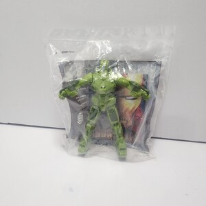 May include: A translucent green Iron Man action figure in a sealed plastic bag. The figure is in a standing pose with arms outstretched. The packaging has a partial view of a graphic with the word "IRON MAN".