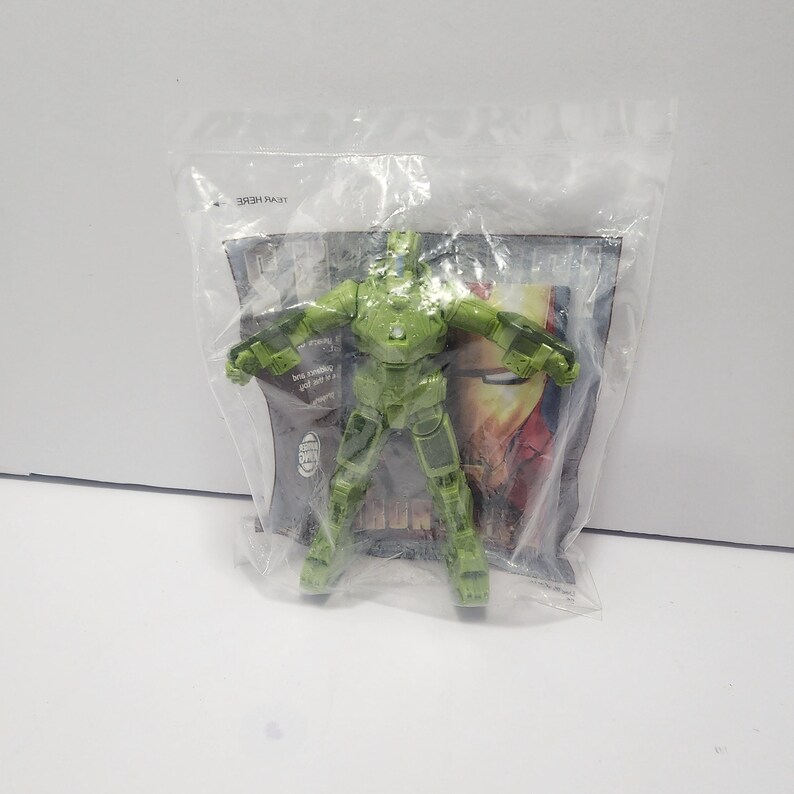 May include: A translucent green Iron Man action figure in a sealed plastic bag. The figure is in a standing pose with arms outstretched. The packaging has a partial view of a graphic with the word "IRON MAN".