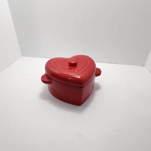 Puede incluir: Una fuente de cerámica roja en forma de corazón con tapa. La fuente tiene asas pequeñas y un pomo en la tapa. La tapa y el borde están decorados con un patrón de puntos. Aproximadamente 15 cm de ancho.