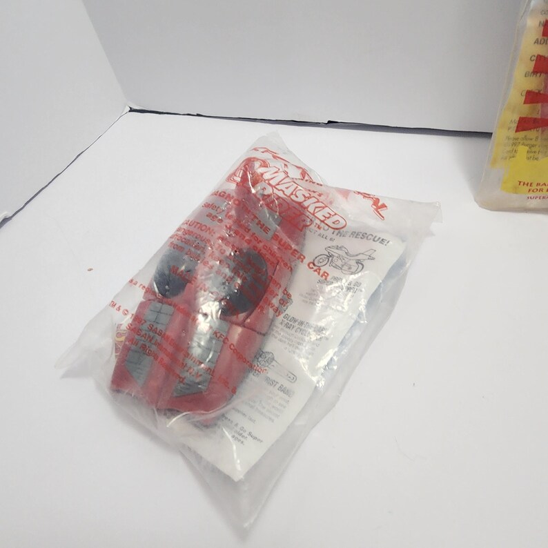 May include: A red toy car is packaged in a clear plastic bag. The bag is printed with the word "MASKED" in red. The car has black wheels and is partially visible. The packaging also includes text and a motorcycle graphic.