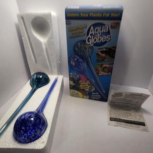 Könnte beinhalten: Zwei handgeblasene Aqua Globes aus Glas zum Gießen von Pflanzen. Einer ist tiefblau mit weißen Sprenkeln, der andere türkis. Sie befinden sich in einem weißen Schaumstoffeinsatz. Eine Schachtel mit Aqua Globes ist ebenfalls sichtbar.