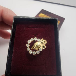 Könnte beinhalten: Ein goldfarbener Ring mit einer Handgeste, umgeben von klaren Edelsteinen. Der Ring wird in einer schwarzen Schachtel mit bordeauxroter Innenausstattung präsentiert, und ein Holzdeckel ist teilweise sichtbar.