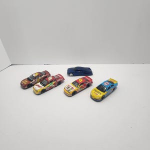 Puede incluir: Una colección de coches de carreras de juguete en varios colores, incluyendo rojo, amarillo, azul y un cupé azul oscuro. Los coches presentan logotipos de patrocinadores y números de carreras. Sobre una superficie blanca.