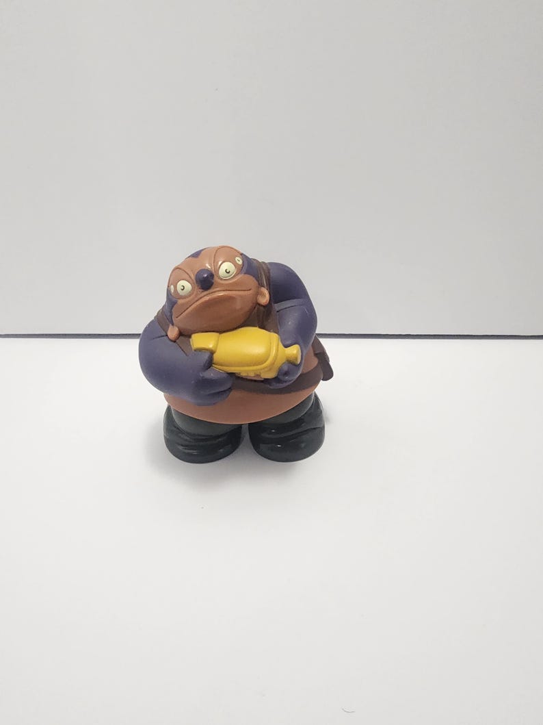 May include: A figurine of a cartoon character with a brown head, purple arms, and brown torso. The character is holding a yellow object and wearing black boots. The figurine is standing on a white surface.