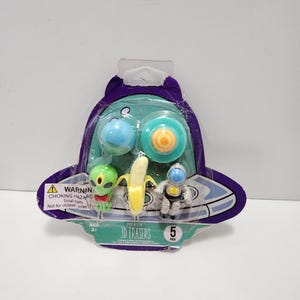 May include: A package of 5 Premiere 3D Erasers featuring a spaceship design. The erasers are shaped like a banana, an alien, an astronaut, and two round objects. The package has a warning label that says "Choking Hazard - Small Parts. Not for children under 3." and an age recommendation of 3+.