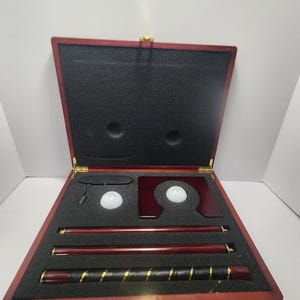 Könnte beinhalten: Ein Golf-Putting-Übungsset in einem Holzkoffer. Das Set enthält einen Putter, zwei Golfbälle und ein Ausrichtungswerkzeug. Der Koffer ist dunkelrot mit goldfarbener Hardware und einem schwarzen Schaumstoff-Innenraum.
