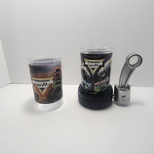 May include: Two clear plastic cups with "Monster Jam" graphics. One cup is on its own, the other is on a black tire base. A silver piston and connecting rod are also present, suggesting a monster truck theme.