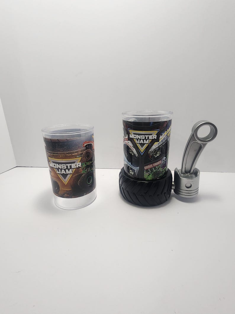 May include: Two clear plastic cups with "Monster Jam" graphics. One cup is on its own, the other is on a black tire base. A silver piston and connecting rod are also present, suggesting a monster truck theme.