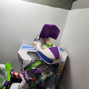 May include: A purple and white toy spaceship with a figure of Buzz Lightyear inside. The spaceship is open and the figure is sitting in the cockpit. The toy is part of a Disney Toy Story set.