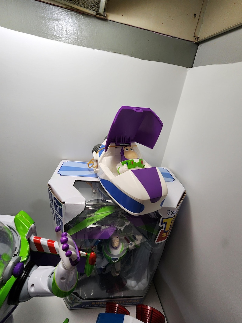 May include: A purple and white toy spaceship with a figure of Buzz Lightyear inside. The spaceship is open and the figure is sitting in the cockpit. The toy is part of a Disney Toy Story set.