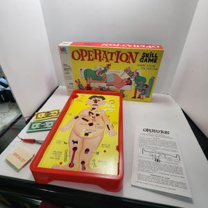 May include: Operation skill game set including a red plastic game board with a cartoon patient, a yellow box with illustrations, a pair of metal tweezers with a red cord, paper money, and a white instruction manual with black text.