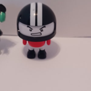 Puede incluir: Una peque&ntilde;a figura de juguete negra y blanca que lleva un casco, una camisa roja y botas negras. La figura tiene una cara de dibujos animados con una expresi&oacute;n de enfado.