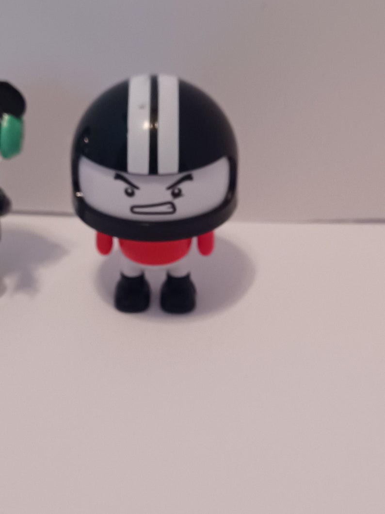 Puede incluir: Una peque&ntilde;a figura de juguete negra y blanca que lleva un casco, una camisa roja y botas negras. La figura tiene una cara de dibujos animados con una expresi&oacute;n de enfado.