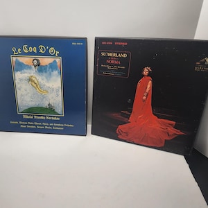 May include: Two vintage vinyl record albums. One album cover is blue with gold lettering and an illustration. The other album cover is black with a woman in a red gown. The albums are labeled "Le Coq D'Or" and "Norma".