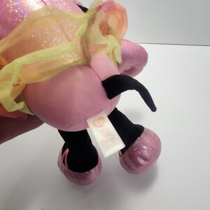 May include: A pink plush toy with black antennae, black legs, and sparkly pink feet. The toy has a yellow and orange tulle accent and a white tag with a heart logo.
