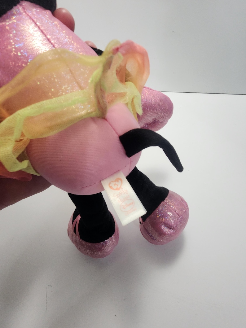 May include: A pink plush toy with black antennae, black legs, and sparkly pink feet. The toy has a yellow and orange tulle accent and a white tag with a heart logo.