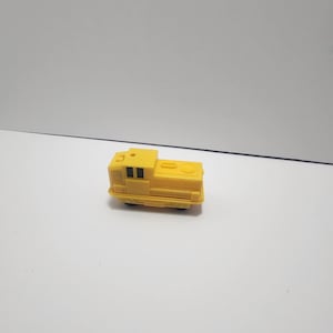 May include: A small, yellow toy train with black windows and wheels. The toy train is a miniature replica of a diesel locomotive, featuring a rectangular body and a rounded top. The toy is made of plastic.