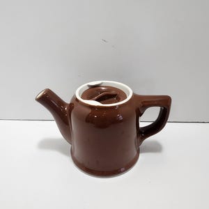 May include: A small, brown ceramic teapot with a white rim around the lid. The teapot has a spout, handle, and a lid with a small knob. The teapot is likely for brewing tea.