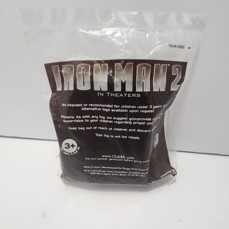 May include: A clear plastic bag with a brown label featuring the text "IRON MAN 2 IN THEATERS". The bag includes text about age recommendations and toy safety. The bag is sealed and has a "TEAR HERE" arrow.