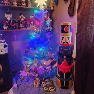 May include: A white artificial Christmas tree with colorful lights and a star on top. A stack of four nutcracker themed gift boxes are next to the tree. The tree is decorated with ornaments and is sitting on a blue and white tree skirt.
