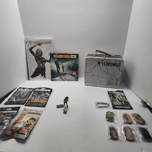 May include: A collection of Walking Dead merchandise, including comic books, a lunchbox with the word "TERMINUS", action figures, and character keychains. The items are arranged on a white surface.