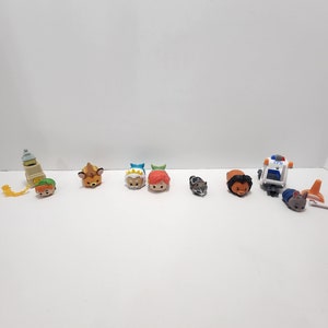 May include: A collection of 10 small plastic figurines, including characters from Disney and Pixar movies. The figurines are in various colors, including green, brown, blue, yellow, orange, and red. The figurines are all in a standing position, and they are arranged in a row on a white surface.