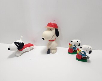 Può includere: Tre figurine di plastica di Snoopy, l'iconico beagle della striscia a fumetti Peanuts. Una figurina raffigura Snoopy con un berretto da baseball rosso e un bastone da baseball. Un'altra figurina mostra Snoopy con una camicia blu e un estintore rosso. La terza figurina mostra Snoopy con un mantello rosso e vola nell'aria.
