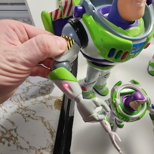 May include: A green and white Buzz Lightyear action figure with a purple helmet and a red light on its arm. The figure is holding a smaller Buzz Lightyear action figure with a purple helmet and a white and green space suit.