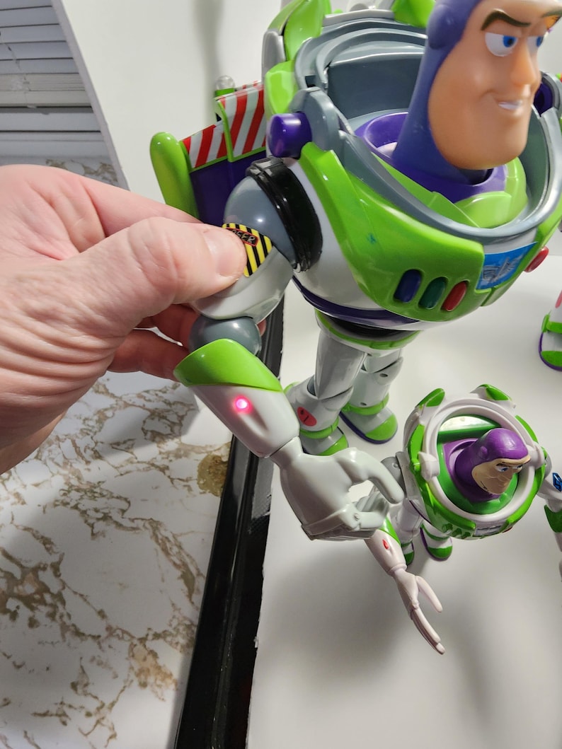 May include: A green and white Buzz Lightyear action figure with a purple helmet and a red light on its arm. The figure is holding a smaller Buzz Lightyear action figure with a purple helmet and a white and green space suit.