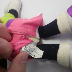 May include: A plush toy featuring a pink skirt, black legs, and white shoes. The toy has a green and purple top. A tag reads "ACOTOYS, INC. WESTBURY, NY. 11590, U.S.A. MADE IN CHINA DISNEY 468KY."
