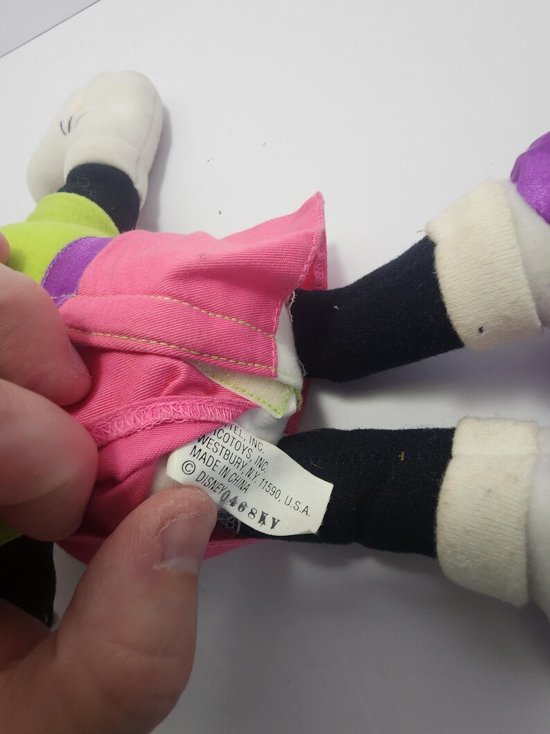 May include: A plush toy featuring a pink skirt, black legs, and white shoes. The toy has a green and purple top. A tag reads "ACOTOYS, INC. WESTBURY, NY. 11590, U.S.A. MADE IN CHINA DISNEY 468KY."
