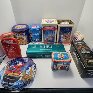 May include: A collection of vintage tin boxes featuring various brands and designs, including Coca-Cola, Snickers, Happy Birthday, Cracker Jack, Pall Mall, Nürnberger Lebkuchen, and Jack Daniel's Tennessee Whiskey. The boxes are in various shapes and sizes, with colorful graphics and logos.