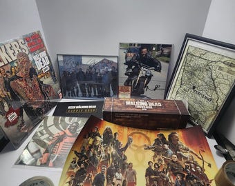 May include: A collection of "The Walking Dead" merchandise, including signed photos, a framed map, and a poster with the text "The Art of the Walking Dead Universe". Also visible are a box and a roll of tape.