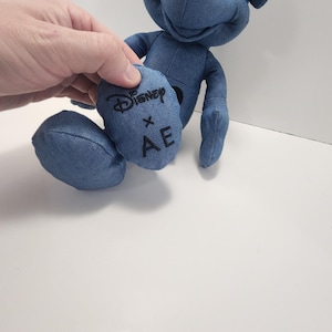 Puede incluir: Un peluche azul con forma de personaje de dibujos animados. El juguete est&aacute; hecho de tela similar al denim y presenta el logotipo de Disney y las letras "AE" en uno de sus pies. El juguete tiene ojos negros y nariz negra.
