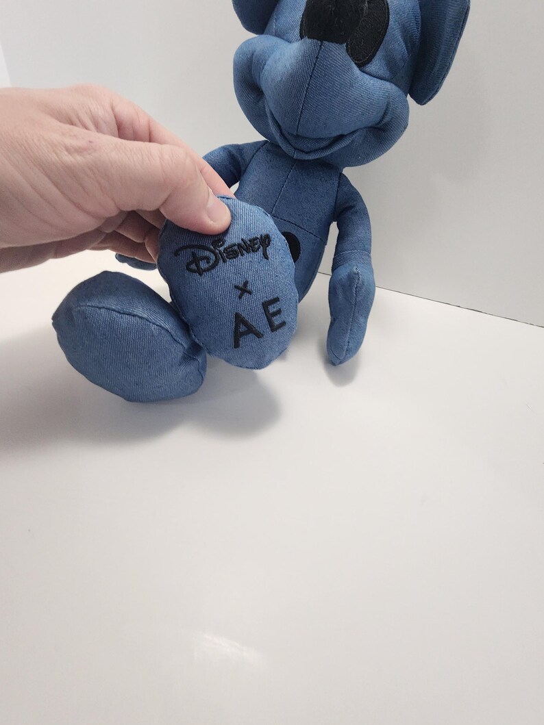 Puede incluir: Un peluche azul con forma de personaje de dibujos animados. El juguete est&aacute; hecho de tela similar al denim y presenta el logotipo de Disney y las letras "AE" en uno de sus pies. El juguete tiene ojos negros y nariz negra.