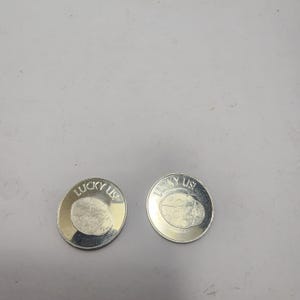 以下が含まれることがあります： てんとう虫のデザインと「LUCKY US」の文字が入った、2つの銀色のコイン。 コインは丸く、銀色の縁取りと、中央に詳細なてんとう虫のイラストが描かれています。