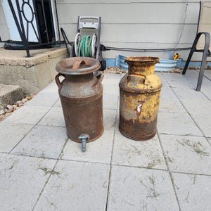 May include: Two vintage metal milk churns. The left churn is dark brown with a metal tap at the base and two side handles. The right churn is yellow with visible rust, featuring a painted deer graphic on the front and two side handles.