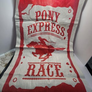 May include: A white and red Pony Express Race banner. The banner features the words "Pony Express" and "Race" in red, along with an illustration of a rider on a horse. The banner is made of a woven material.