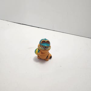 May include: Orange Garfield figurine wearing a blue and yellow hat and holding a yellow and blue ball. The figurine has black stripes and a smiling face. The figurine is sitting on a white surface.