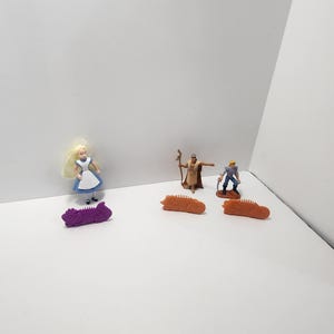 Può includere: Una collezione di piccole statuette e giocattoli, tra cui una bambola con un vestito blu, una figura con un bastone e una figura con un piccone. Sono inclusi anche tre alligatori giocattolo arancioni e uno viola.