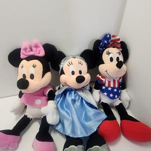 May include: Three plush Mickey Mouse toys. One is wearing a pink dress and bow, one is wearing a blue dress and a blue bow, and one is wearing a red, white and blue outfit with a red bow.