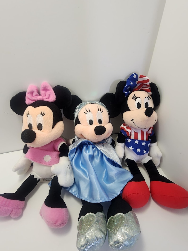 May include: Three plush Mickey Mouse toys. One is wearing a pink dress and bow, one is wearing a blue dress and a blue bow, and one is wearing a red, white and blue outfit with a red bow.