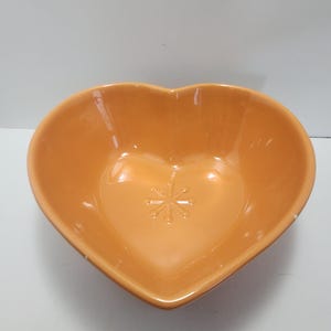 May include: An orange heart-shaped ceramic bowl. The bowl has a smooth, glossy finish and a decorative floral design in the center. The bowl is empty and sits on a white surface.