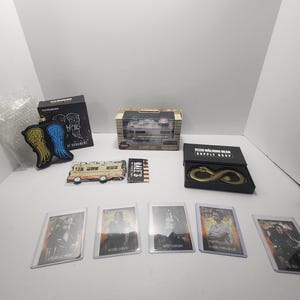 May include: A collection of merchandise featuring items from "The Walking Dead." Includes trading cards, a gold-colored infinity symbol, a miniature RV, and an ornament with angel wings. The items are displayed on a white surface.