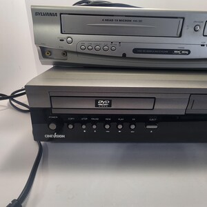 Może przedstawiać: Srebrny odtwarzacz VCR Sylvania ułożony na czarno-srebrnym odtwarzaczu DVD Cinevision. VCR ma przyciski zasilania, przewijania i odtwarzania. Odtwarzacz DVD ma przyciski zasilania, kopiowania, zatrzymywania, pauzy, przewijania, odtwarzania, szybkiego przewijania do przodu i wysuwania.