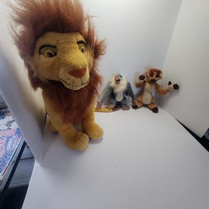 May include: Three plush toys from the Disney movie The Lion King. The toys are Simba, Rafiki, and Timon. Simba is a large, brown and yellow lion with a mane of brown fur. Rafiki is a small, gray monkey with a purple and blue shirt. Timon is a small, brown meerkat with a yellow shirt.