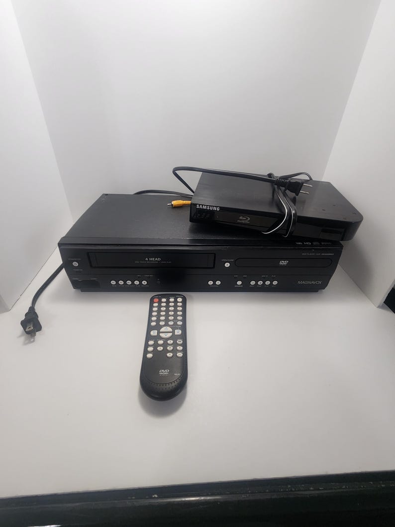 K&ouml;nnte beinhalten: Ein schwarzer DVD/VHS-Kombinations-Player mit einer Fernbedienung. Der Player verf&uuml;gt &uuml;ber ein 4-Kopf-System und ein DVD/VHS-Logo. Der Player steht auf einem schwarzen Samsung Blu-ray-Player.