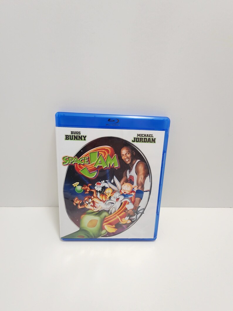 Puede incluir: Una caja de Blu-ray azul con una imagen de Bugs Bunny, Daffy Duck y Michael Jordan jugando baloncesto sobre un fondo espacial. El texto "Space Jam" est&aacute; en el centro de la imagen. Los textos "Bugs Bunny" y "Michael Jordan" est&aacute;n en las esquinas superiores de la caja.