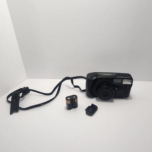 May include: A black Maxima Zoom Pro 70 film camera with a lens, flash, and strap. Includes a battery pack and a lens cap. The camera is on a white surface.
