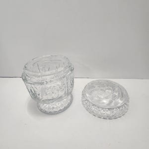 May include: A clear glass jar with a lid. The jar has a decorative pattern on the sides and the lid has an embossed design. The jar is open, and the lid is next to it. The jar sits on a white surface.